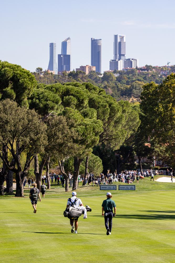 Un golfista y su caddie caminan por el Recorrido Negro de golf del Real Club de Campo Villa de Madrid.on las Torres de Madrid al fondo.